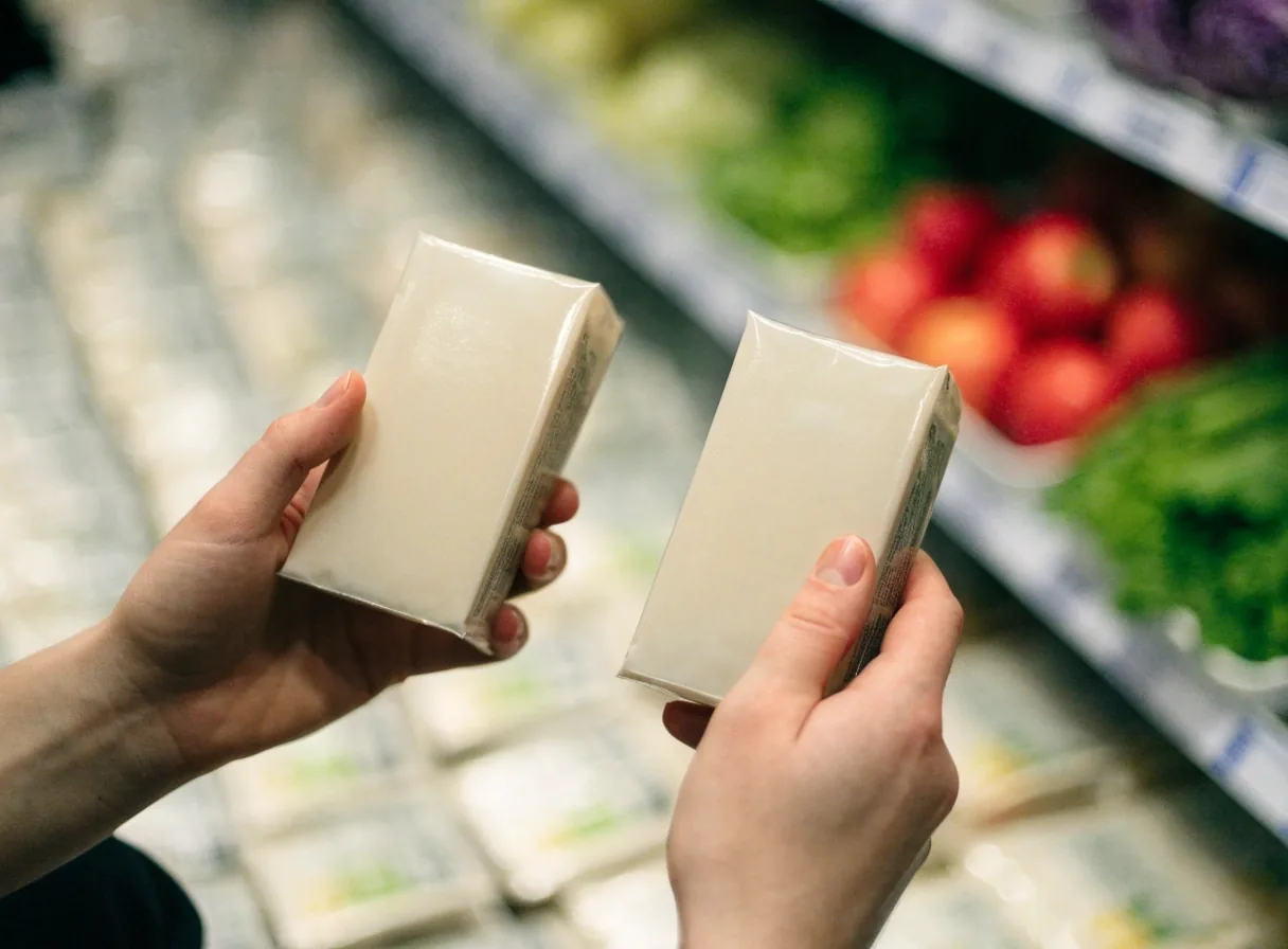 Two hands comparing two unbranded packaged items on a store shelf with fresh produce behind